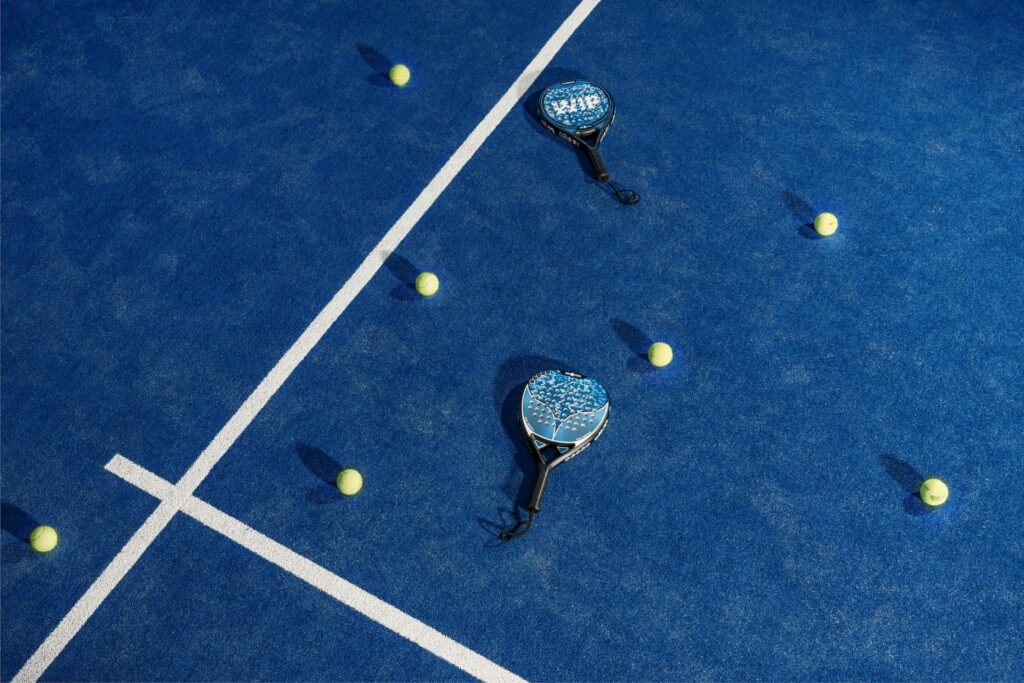 Twee padelrackets en zes gele ballen verspreid op blauwe padelbaan