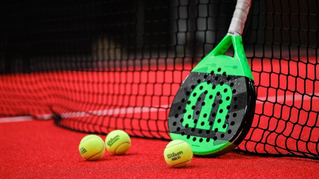 Groen-zwarte padelracket en Wilson padelballen op rode baan naast het net