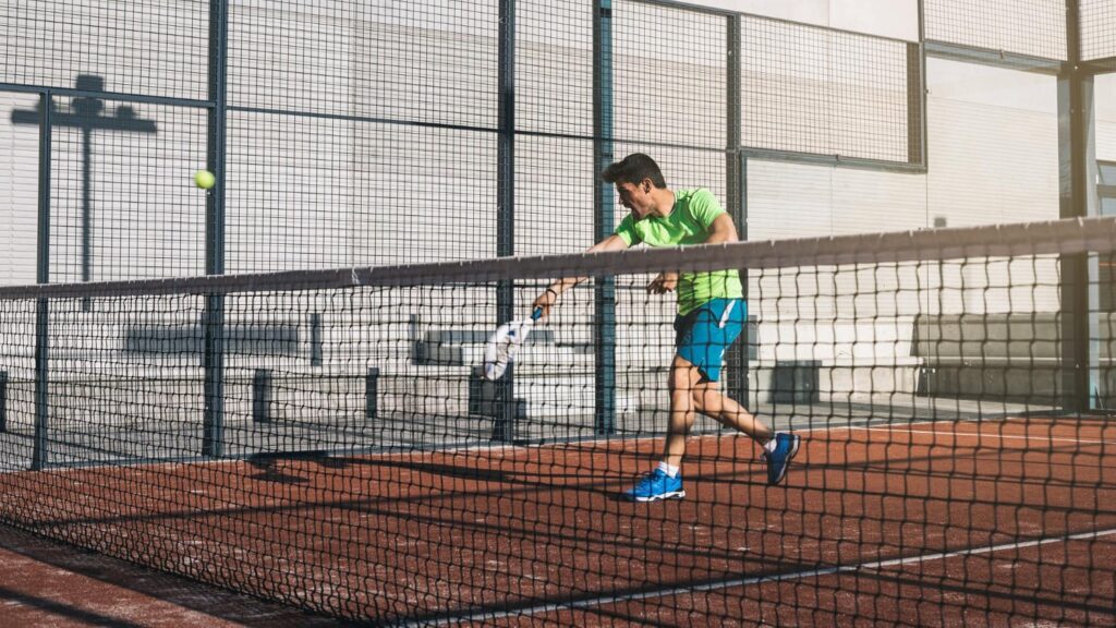 Man in groene sportkleding slaat met een padel racket een bal op een buitenbaan, tijdens een intensieve padelwedstrijd.
