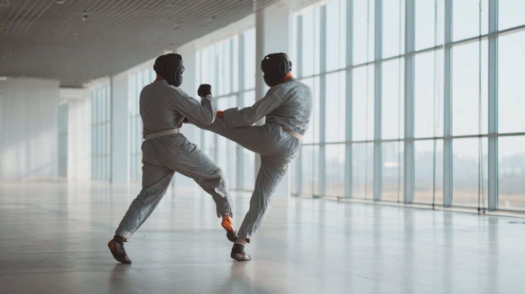 Twee kickboksers in beschermende pakken en hoofdbescherming oefenen een trapaanval tijdens een sparringssessie in een lichte sportruimte