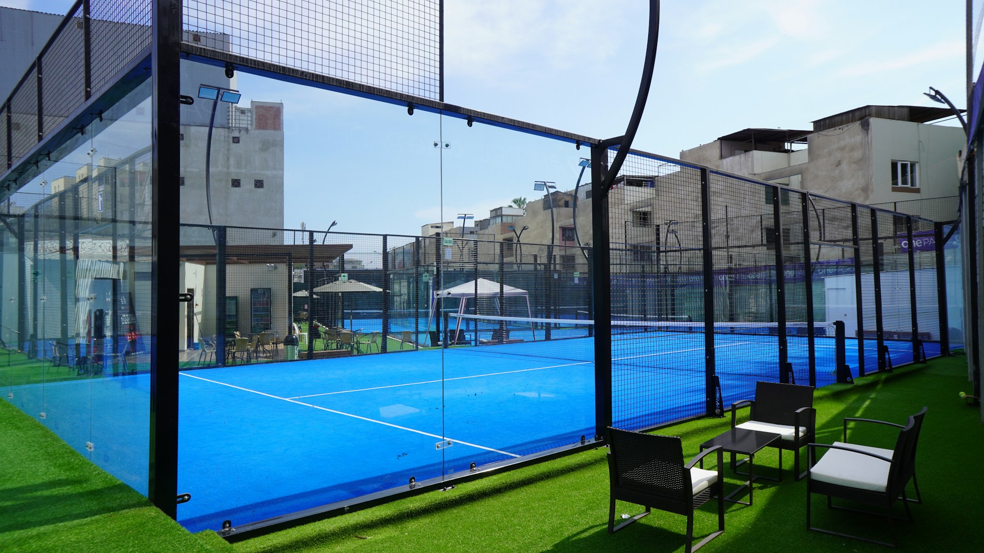 Blauwe padelbaan met glazen wanden en een loungehoek met rieten stoelen op kunstgras, sfeer die je in een padelclubhuis kunt inrichten
